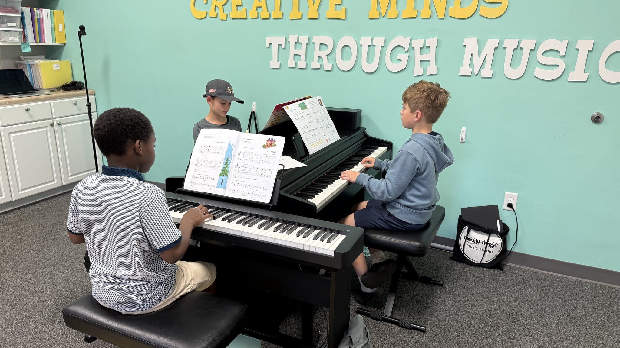 Three children playing keyboards together in a group piano class under the Creative Minds Through Music sign